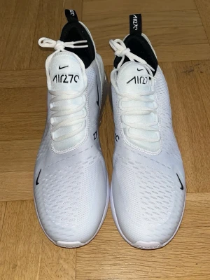 Nike Air Max 270 vita sneakers - Nike Air Max 270 sneakers i vitt med svarta detaljer och synlig luftbubbla i hälen. Ovandelen är i mesh för bra ventilation och har tydliga Air 270-loggor på plösen och hälen. Snygg och sportig design med klassisk Nike Swoosh på sidan.