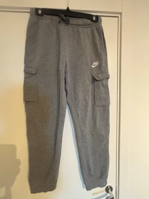 Grå cargobyxor från Nike - Snygga grå cargobyxor från Nike med resår i midjan och dragsko. Byxorna har två stora cargofickor på sidorna och ribbade benslut för en avslappnad look. Tillverkade i mjukt sweatshirtmaterial som är skönt att ha på sig. Nike-logga broderad på vänster ben.