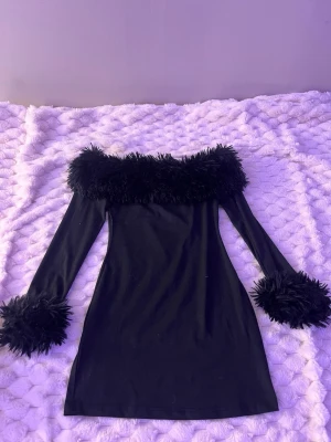 Svart offshoulder klänning med fjädrar - Trendig svart offshoulder klänning med långa ärmar och fluffiga fjäderdetaljer vid halsringning och ärmslut. Kort modell som ger en cool och unik look, perfekt för dig som vill sticka ut. Materialet känns mjukt och följsamt.