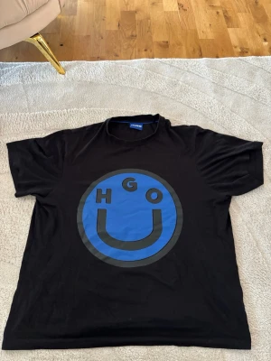 Svart t-shirt med blå smiley från Hugo - Säljer en svart t-shirt från Hugo med en stor blå smiley och bokstäverna HGO på bröstet. T-shirten har rund hals, korta ärmar och är i mjuk bomull. Perfekt för dig som gillar streetwear och vill sticka ut med en cool grafisk detalj. Den passar L för den är baggy 