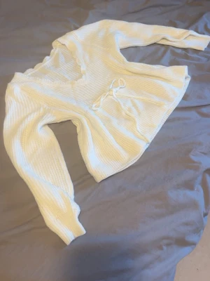 Vit stickad topp med knytband - Säljer en vit stickad topp med v-ringning och dekorativt knytband i midjan. Tröjan har långa ärmar och ett fint ribbstickat mönster. Perfekt för dig som gillar en enkel och stilren look. Materialet känns mjukt och bekvämt.