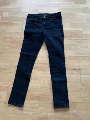 Nudie Jeans - Säljer ett par svarta slim fit jeans från Nudie med klassisk femficksdesign och raka ben. Jeansen har snygga detaljer och är tillverkade i bomull med stretch för extra komfort. jätte bra för dig som gillar en riktigt stilren och modern look.