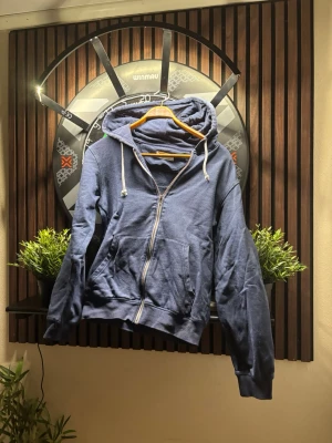 Blå zip hoodie från Pier One - Mysig blå hoodie från Pier One med dragkedja framtill, två stora fickor och vit dragsko i huvan. Tillverkad i mjukt material med borstad insida för extra komfort. Perfekt för chill dagar eller när du vill ha en avslappnad stil.