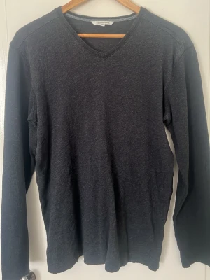 Mörkgrå V-ringad tröja Calvin Klein - Mörkgrå långärmad tröja från Calvin Klein Jeans med V-ringning. Tillverkad i mjukt material som känns skönt mot huden. Enkel och stilren design som passar perfekt till jeans eller chinos.