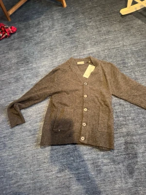 Brun kofta med knappar och fickor - Mysig brun kofta med v-ringning och stora knappar framtill. Två rymliga fickor på framsidan och ribbade muddar vid ärmslut och nederkant. Perfekt för lager på lager och enkel att matcha med olika outfits.