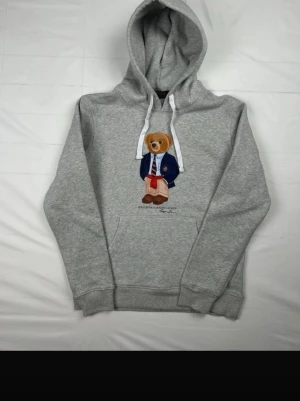 Grå hoodie med björnmotiv från Polo Ralph Lauren - Snygg grå hoodie från Polo Ralph Lauren med det klassiska björnmotivet på bröstet. Hoodien har vit dragsko i huvan och en stor magficka. Perfekt för en chill och avslappnad stil. Materialet är mjukt och skönt, troligen bomullsblandning.