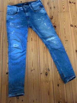 Blå slitna jeans från Replay - Säljer ett par blå jeans från Ralph Lauren med slitningar och distressed detaljer på både fram- och baksida. Jeansen har klassisk femficksmodell och smal passform. Perfekta för en avslappnad och trendig look.