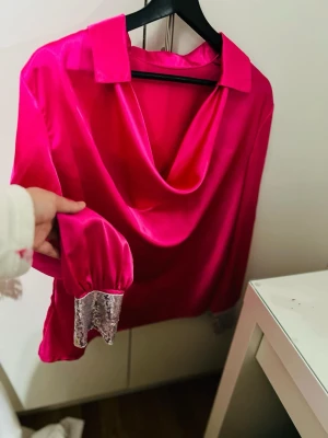 Fuchsia satinblus med paljettmudd - Snygg fuchsiafärgad satinblus med draperad halsringning och krage. Långa ärmar med glittrande paljettmuddar som ger en extra touch. Perfekt för dig som gillar att sticka ut och vill ha något unikt i garderoben.