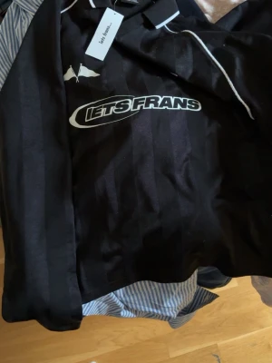 Svart t-shirt från Iets Frans - Cool svart t-shirt från Iets Frans med vit logotyp på bröstet och diskreta ränder i tyget. T-shirten har en klassisk krage och vit piping längs axlarna. Materialet är mjukt och känns bekvämt mot huden. Perfekt för dig som vill ha en unik och trendig look.