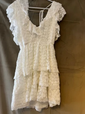 Vit spetsklänning med volanger -   Vit fin klänning med volangdetaljer och v-ringning, knytband i ryggen. Nästan helt som ny, använd 1 gång inga fläckar. 