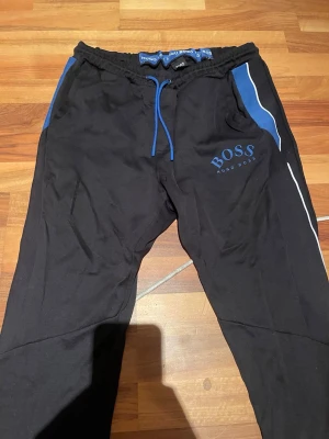 Svarta mjukisbyxor från Hugo Boss - Snygga svarta mjukisbyxor från Hugo Boss med blå detaljer och vita ränder längs benen. Byxorna har dragsko i midjan och fickor på sidorna. Boss-logga tryckt på vänster lår. Perfekta för en avslappnad och sportig stil. OBS: Lite slitna