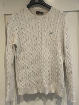 Vit kabelstickad tröja Morris - Kabelstickad tröja i offwhite från Morris med rund halsringning och diskret broderad logga på bröstet. Tröjan har ribbade muddar vid ärmslut och nederkant. Perfekt för dig som gillar klassisk och stilren look.