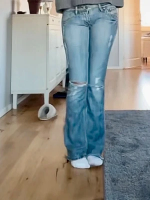 Blå slitna bootcut jeans från diesel - Säljer ett par ljusblå bootcut jeans med slitningar och hål vid knäna. Jeansen har klassisk femficksmodell och låg midja. Perfekta för en avslappnad och trendig look.