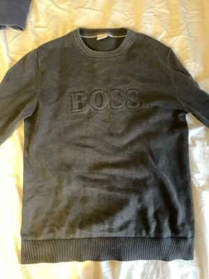Svart sweatshirt från Hugo Boss - Snygg svart sweatshirt från Hugo Boss med broderad logga på bröstet. Tröjan har rund halsringning, ribbade muddar vid ärmslut och nederkant samt en klassisk passform. Perfekt för en stilren och avslappnad look.