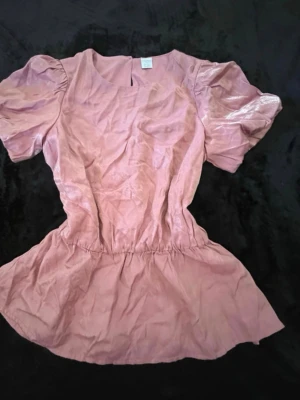 Rosa topp med puffärmar - Söt rosa topp med puffiga kortärmar och markerad midja. Toppen har en lätt glansig finish och en feminin, utsvängd nederdel. Perfekt för dig som gillar romantisk och trendig stil. Egentligen storlek 170/15år. Men passar bra på kvinna/dam i storlek XS-liten M. Har bundle rabatter! 