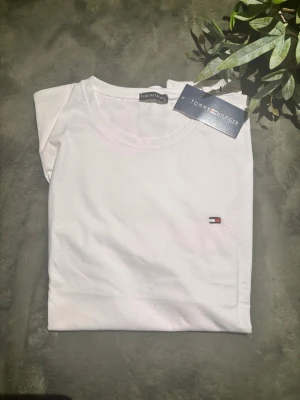Vit t-shirt från Tommy Hilfiger - Klassisk vit t-shirt från Tommy Hilfiger med rund halsringning och liten broderad logga på bröstet. Tillverkad i mjuk bomull för en skön känsla. Enkel och stilren design som passar till det mesta.