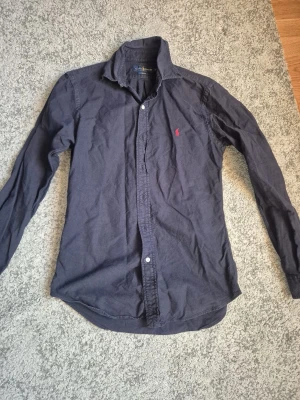 Mörkblå skjorta från Ralph Lauren - Klassisk mörkblå skjorta från Ralph Lauren i slim fit-modell. Skjortan har långa ärmar, knappar framtill och den ikoniska röda loggan broderad på bröstet. Tillverkad i mjuk bomull som känns skön mot huden. Perfekt för en clean och stilren look.