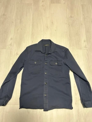 Mörkblå overshirt från Gabba - Stilren mörkblå overshirt från Gabba med raka linjer och två bröstfickor med lock och knapp. Klassisk krage och knäppning framtill. Perfekt lager på lager-plagg i ett mjukt bomullstyg. Storlek M