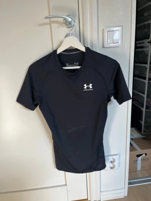 Svart kompressionströja Under Armour - Säljer en svart kompressionströja från Under Armour med korta ärmar och rund hals. Tröjan har logga på bröstet och i nacken, och är tillverkad i ett stretchigt funktionsmaterial som passar perfekt till träning och sport.