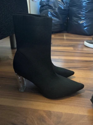 Svarta stretchiga stövletter med klack - Säljer ett par svarta stövletter med stickad, stretchig ovandel och spetsig tå. Skorna har en transparent blockklack som ger en modern touch. Materialet är textil med diskret fiskbensmönster. Perfekta för dig som vill ha något trendigt och bekvämt.