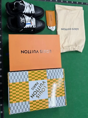 Svarta Louis Vuitton sneakers - Snygga svarta sneakers från Louis Vuitton med vita snören och klassisk LV-logga på plösen. Skorna har en sportig siluett och detaljer i vitt och orange. Kommer med originalkartong, dustbag och extra skosnören.