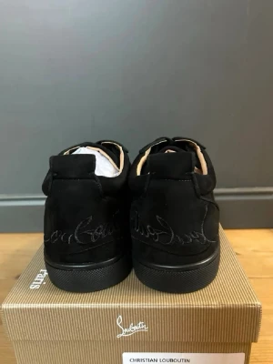 Svarta sneakers från Christian Louboutin - Snygga svarta sneakers från Christian Louboutin i mocka med klassisk röd sula och broderad logga på hälen. Skorna har svarta skosnören och låg profil som ger en clean och exklusiv look. Perfekta för dig som gillar lyxiga detaljer och streetstyle.