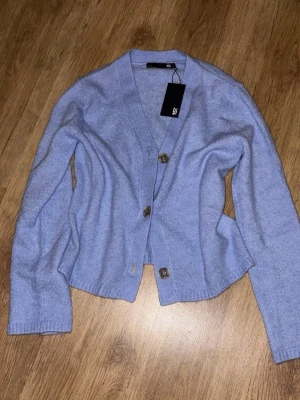 Ljuslila kofta från Lager 157 - Säljer en ljuslila kofta från Lager 157 med v-ringning och stora knappar framtill. Koftan har långa ärmar och ribbade muddar vid ärmslut och nederkant. Perfekt att slänga över en t-shirt för en chill look.