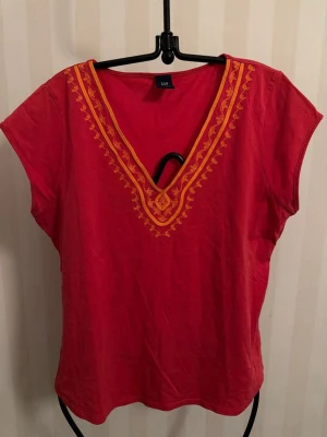 Röd v-ringad topp från GAP - Snygg röd topp från GAP med gul broderad detalj runt den djupa v-ringningen. Kortärmad modell i mjuk bomull, perfekt för en avslappnad och färgstark look. Passar till jeans eller kjol för en cool vardagsstil.