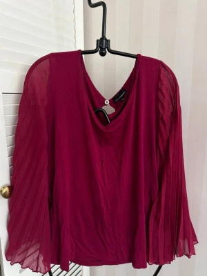 Vinröd plisserad blus med vida ärmar - Snygg vinröd blus från Kapp Ahl med plisserade, transparenta långärmar och rund halsringning. Blusen har en lös passform och knappdetalj bak i nacken. Tillverkad i ett lätt och luftigt material som ger ett elegant intryck.