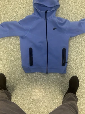 Blå Nike hoodie med dragkedja - Snygg blå hoodie från Nike med hel dragkedja framtill och två svarta dragkedjeförsedda fickor. Klassisk Nike-logga på bröstet och stor huva. Tillverkad i mjukt material som känns skönt mot huden. Perfekt för en avslappnad streetwear-look.