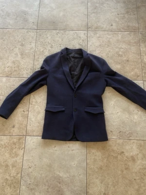 Mörkblå klassisk kavaj - Snygg mörkblå kavaj med två knappar framtill och klassisk passform. Kavajen har två fickor med lock, bröstficka och slits bak. Fodrad insida och stilren design som passar till många olika looks.