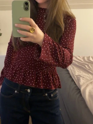 Vinröd prickig blus med smock - Vinröd blus med vita prickar och smockad midja som ger en snygg passform. Blusen har långa ärmar och lätt utsvängd nederkant. Materialet ser ut att vara mjukt och stretchigt, perfekt för att skapa en cool och avslappnad look.
