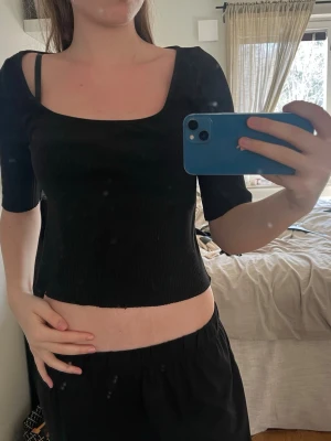 Svart ribbad croppad topp - Säljer en svart ribbad topp med croppad passform och fyrkantig ringning. Toppen har korta ärmar och är tillverkad i ett stretchigt material som sitter snyggt på kroppen. Perfekt att styla med både jeans och kjol.