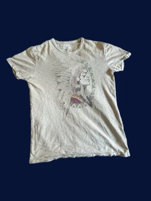 Beige Denim & Supply t-shirt med tryck  - Säljer en beige t-shirt med ett stort tryck av en person med fjäderprydd huvudbonad på bröstet. T-shirten har rund hals, korta ärmar och en avslappnad passform. Perfekt till jeans eller shorts för en chill look.