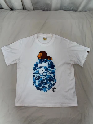 Vit BAPE t-shirt med blått camomönster - Säljer en vit t-shirt från BAPE med det klassiska blå camo-apansikte trycket på bröstet och en liten brun apa ovanpå. T-shirten har korta ärmar och är gjord i mjuk bomull. Liten BAPE-logga på ärmen för extra detalj.