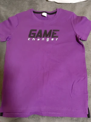 Helt ny. Lila t-shirt GAME CHANGER Lindex - Snygg lila t-shirt från Lindex med trycket 'GAME changer' i svart och vitt på bröstet. Klassisk rund halsringning och korta ärmar. Tillverkad i mjuk bomull som är skön att ha på sig. Perfekt för dig som vill sticka ut lite extra.