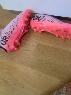 Nike CR7 rosa fotbollsskor - Säljer ett par snygga fotbollsskor från Nike CR7-kollektionen i rosa med svarta detaljer och CR7-logga på hälen. Skorna har låg profil, snörning och dobbar för bra grepp på planen. Tillverkade i syntetmaterial för lätt känsla och bra bollkontroll.