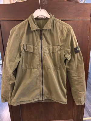 Olivgrön overshirt från Stone Island - Snygg olivgrön overshirt från Stone Island med dragkedja framtill och två bröstfickor med lock. Klassisk krage och ikonisk patch på vänster ärm. Tillverkad i bomull för en skön och avslappnad look. Perfekt lager på lager-plagg för streetwear-stilen.