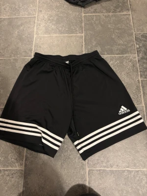 Svarta Adidas shorts med vita ränder - Snygga svarta shorts från Adidas med klassiska vita ränder längst ner på benen och Adidas-logga på vänster sida. Shortsen har snörning i midjan och är tillverkade i ett lätt och bekvämt syntetmaterial, perfekta för sport eller chill.