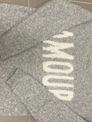 Grå stickad tröja med text - Mysig grå stickad tröja med fluffig vit text 'AMOUR' framtill. Tröjan har långa ärmar och ribbade muddar vid ärmslut och nederkant. Perfekt för dig som gillar en avslappnad och trendig stil.