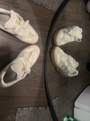 Vita Adidas sneakers - Snygga vita Adidas sneakers med klassiska tre ränder på sidan och gulddetaljer på plösen. Skorna har en låg profil och en ljusbrun gummisula som ger retrovibbar. Tillverkade i läder och textil för en clean look. Perfekta för dig som gillar stilrena och tidlösa sneakers.