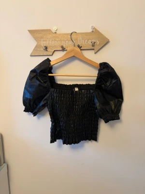 Svart skin puffärmad topp  - Trendig svart topp med puffärmar i skin material. Fyrkantig urringning och kort, croppad passform (inte jätte). Perfekt för en utekväll.