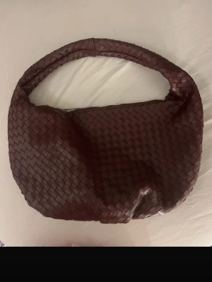 Vinröd flätad axelväska i läder - Säljer en vinröd handväska med flätad design och rundad form. Väskan har ett bredare axelband och dragkedja upptill. Tillverkad i läderliknande material och har en trendig, modern look som sticker ut.