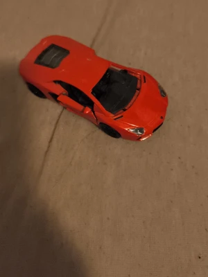 Röd leksaksbil sportbil - Säljer en röd leksaksbil i sportbilsmodell med svarta detaljer och öppningsbara dörrar. Bilen är tillverkad i metall och plast och har realistiska former och detaljer som liknar en Lamborghini. Perfekt för dig som gillar bilar eller samlar på modellbilar.