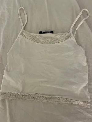 Vit spetsdetaljerad linnetopp FB Sister - Söt vit linnetopp från FB Sister i storlek XS. Toppen har tunna axelband och fina spetsdetaljer både vid halsringningen och nederkanten. Perfekt att styla med jeans eller kjol för en fräsch look.