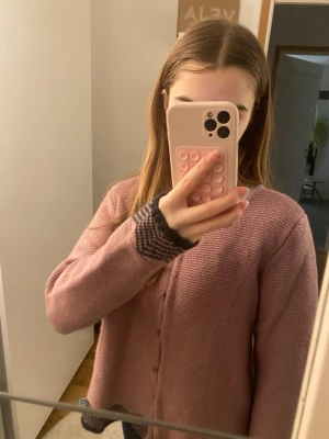 Rosa kofta med knappar och mönster - Mysig rosa kofta med knäppning framtill och dekorativt mörkgrått mönster vid ärmslut och nederkant. Koftan har en rak passform och är stickad i ett mjukt material, perfekt för lager-på-lager-stil.