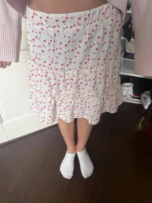 Vit volangkjol med körsbärsmönster - Supersöt vit kortkjol med små röda körsbär och volangdetaljer. Kjolen har resår i midjan och ett luftigt, flowy fall som ger en romantisk vibe. Perfekt för varma dagar och enkel att styla med en stickad tröja eller crop top.