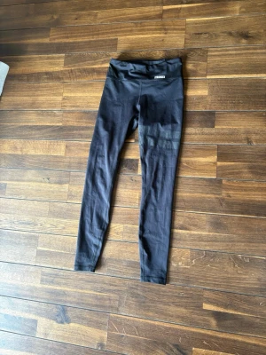 Svarta leggings från Stronger - Säljer ett par svarta leggings från Stronger med hög midja och stretchigt material. Perfekta för träning eller chill, med diskret logga framtill i midjan. Snygg passform som sitter tajt längs benen.