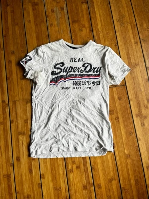 Vit Superdry t-shirt med tryck - Vit t-shirt från Superdry med stort svart och rött tryck på bröstet och japanska tecken. Klassisk rund hals och korta ärmar. Materialet är mjuk bomull och det finns även ett litet tryck på ena ärmen. Perfekt för dig som gillar streetstyle.