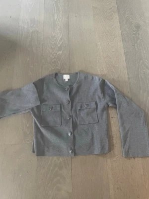 Mörkgrå kofta med fickor - Stilren mörkgrå kofta med rund halsringning och knäppning framtill. Två stora bröstfickor med lock och knappdetaljer ger en trendig look. Perfekt att slänga över en t-shirt för en avslappnad men snygg outfit.